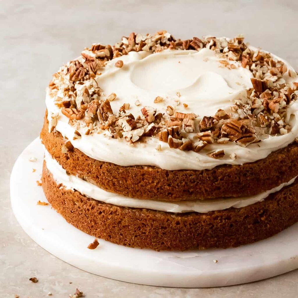 Overhead view of a freshly baked carrot cake on a white dish, topped with a generous layer of cream cheese frosting and a sprinkle of chopped nuts, ready to be served.